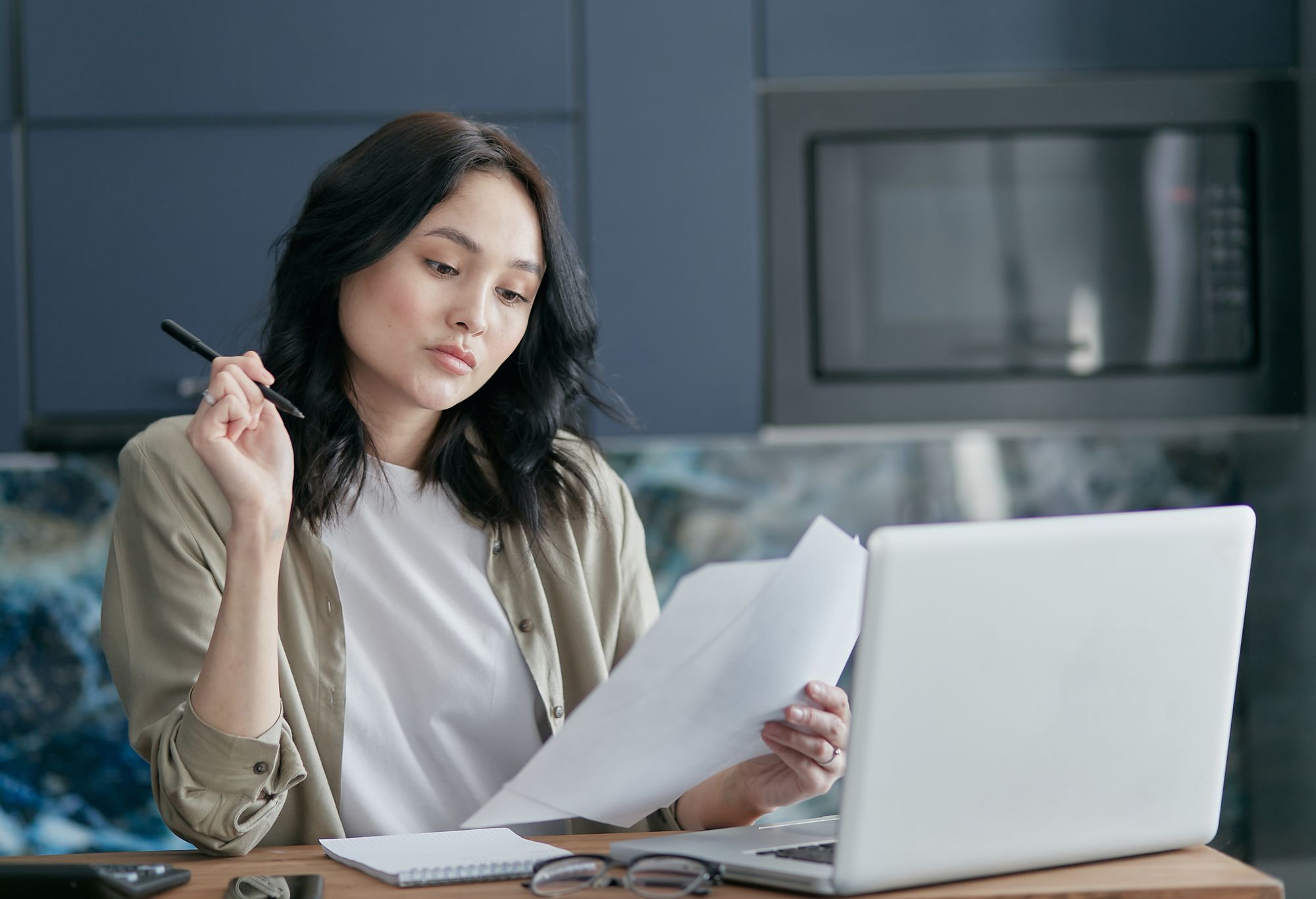 Business owner reading loan paperwork at her kitchen bench at home, laptop open beside her